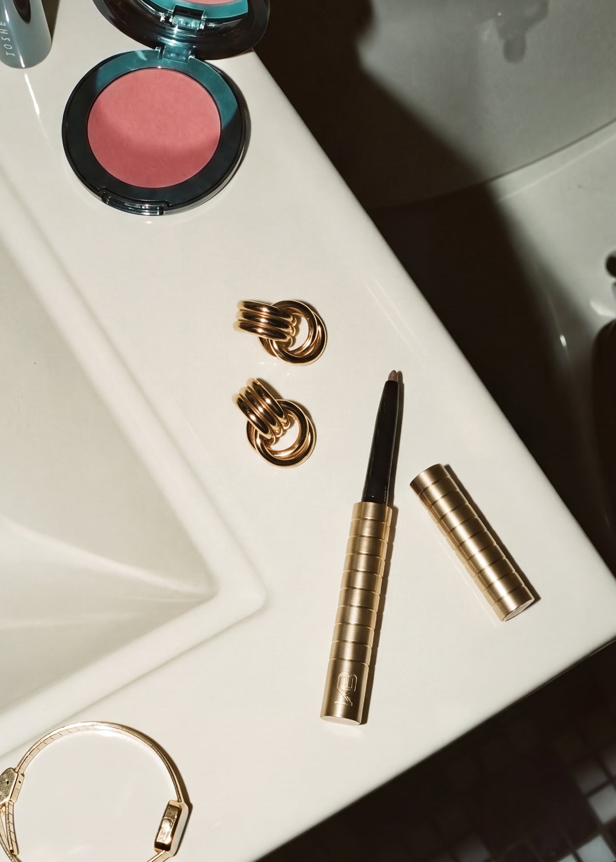 Gold-toned Karen Betts pen and casing on a bathroom basin beside gold earrings and a pink blusher