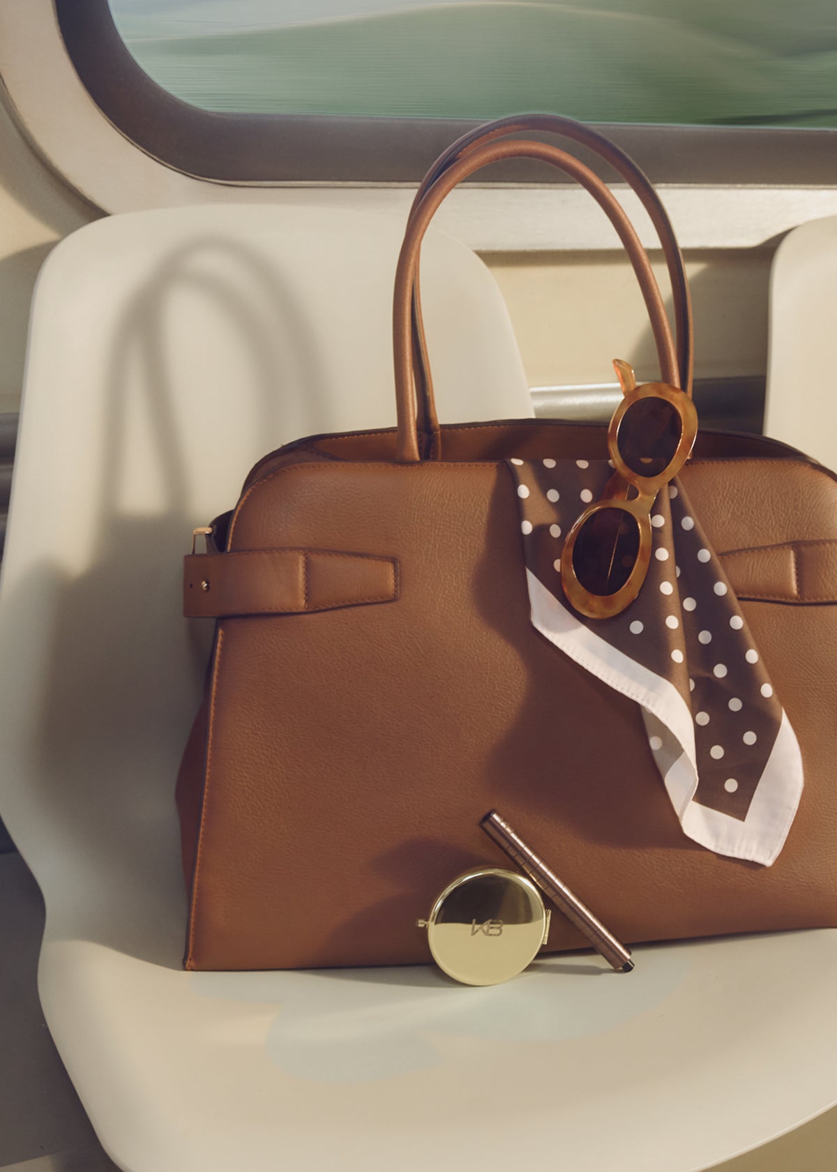 Brown leather handbag on a car seat with sunglasses, a KB compact mirror and a Karen Betts pen