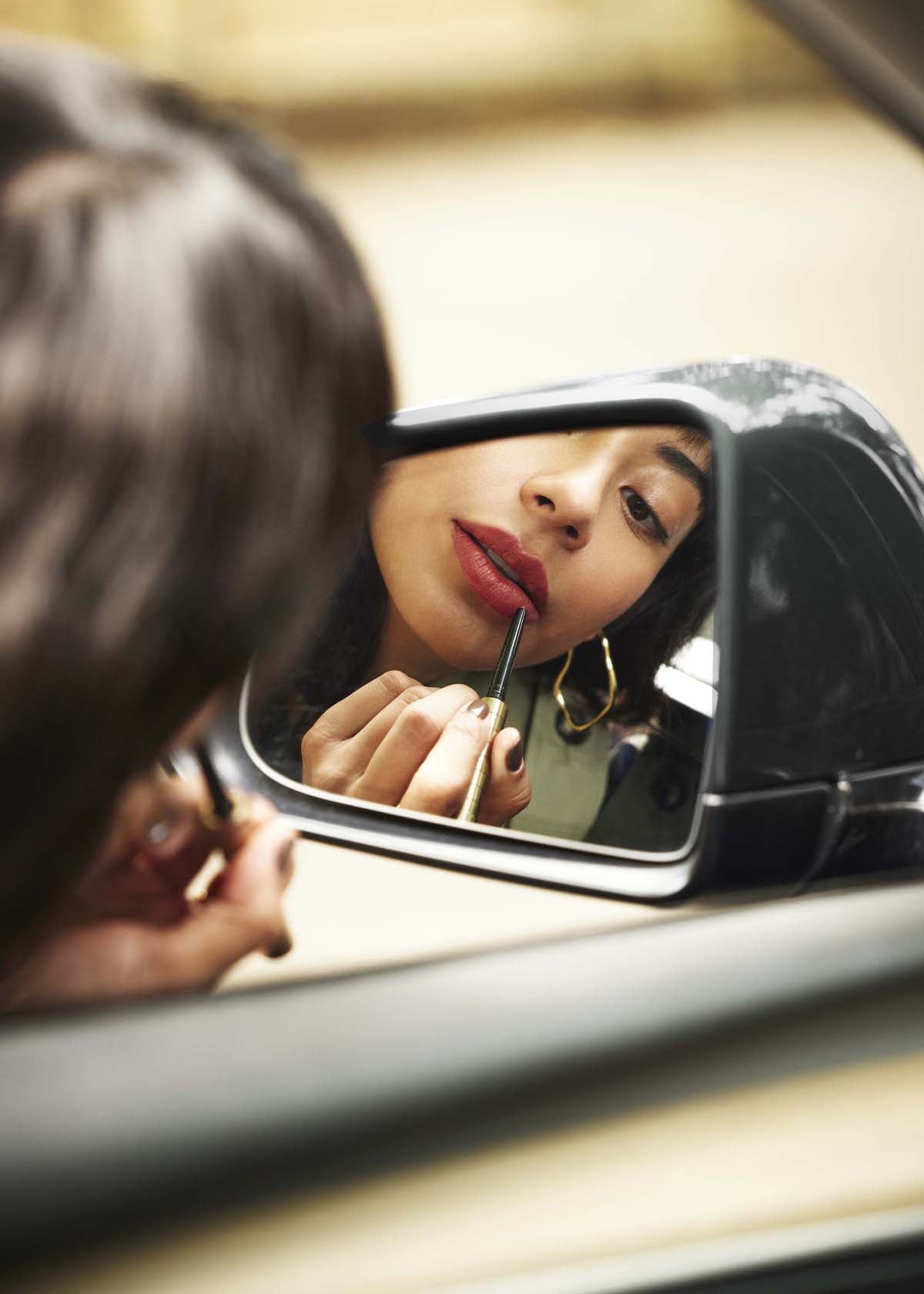 Woman lining her lips in red with the Karen Betts pen in a car wing mirror