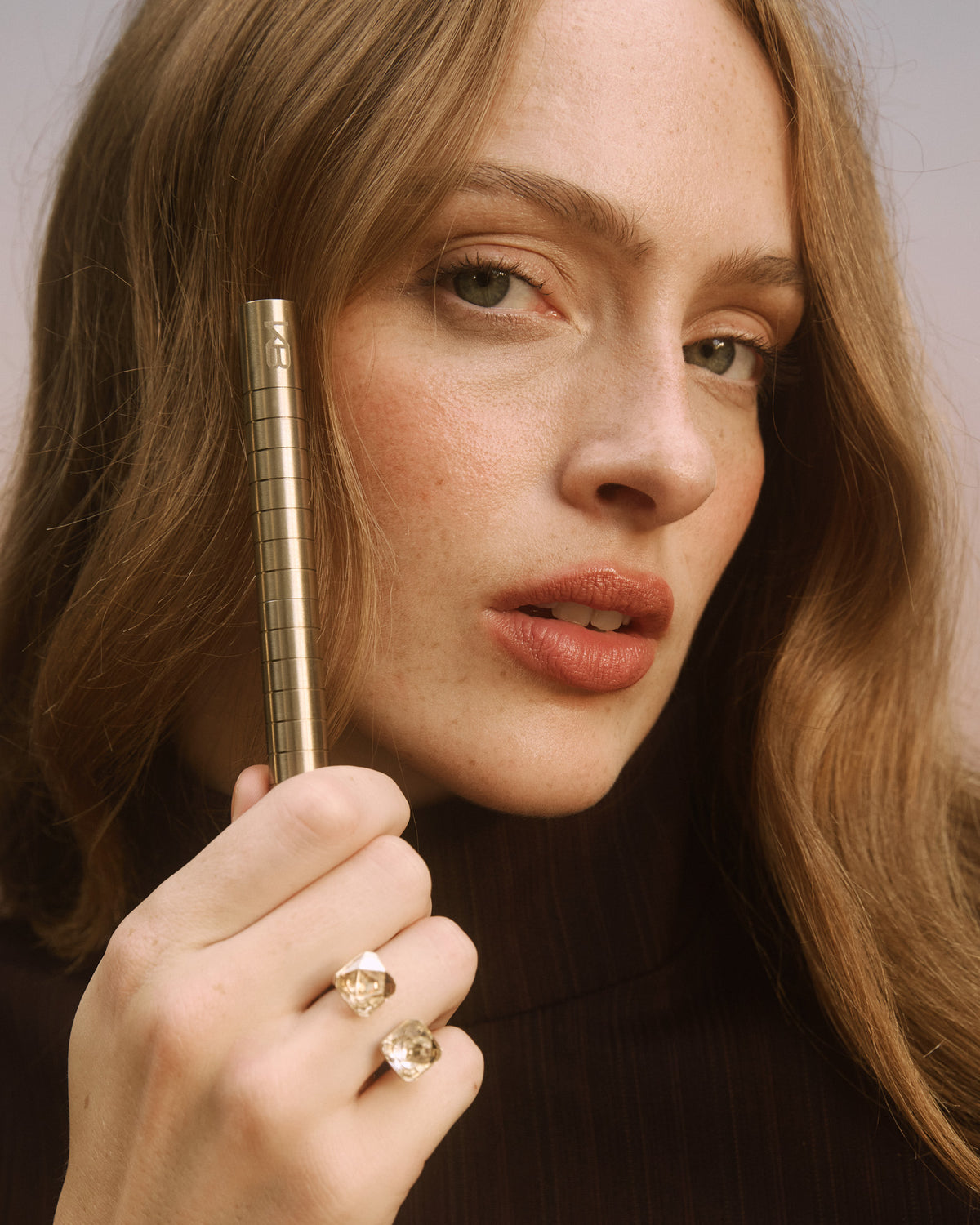 Woman holding a gold-toned Karen Betts pen to her cheek, wearing a soft terracotta lip